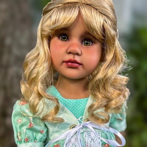 May include: A doll with long, wavy blonde hair, green eyes, and freckles. The doll wears a light green dress featuring a small floral pattern, a white lace trim down the center, and a white ribbon tied in a bow at the chest with gold eyelets.