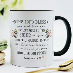 May include: White ceramic mug with a black handle and rim, featuring the text "THE LORD BLESS" and a floral design. The mug is next to two macarons and a gold spoon. The text is a quote from Numbers 6:24-26.