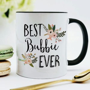 May include: White ceramic mug with a black rim and handle. The mug has a floral design and the text "BEST Bubbie EVER" in black.