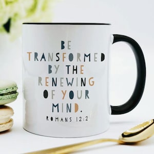 May include: White ceramic mug with a black rim and handle. The mug has a quote printed on it in colorful text: "Be transformed by the renewing of your mind. Romans 12:2".