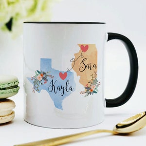 May include: White ceramic coffee mug with a black rim and handle. The mug features a watercolor design of the states of Texas and Illinois with the names "Kayla" and "Sara" written on each state. The states are connected by a line with hearts.