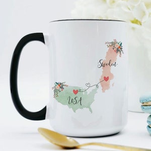 May include: White ceramic mug with a black handle. The mug features a watercolor illustration of the USA and Sweden connected by a line with hearts. The text "USA" and "Sweden" are written on the mug.