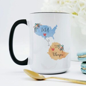 May include: White ceramic mug with a black handle. The mug features a watercolor illustration of the USA and Ethiopia connected by a line of hearts. The text "USA" and "Ethiopia" are written on the map.