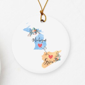 May include: A white ceramic ornament with a blue and red watercolor map of Michigan and Syria connected by a red heart string. The ornament reads "Michigan" and "Syria" with floral accents.