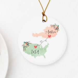May include: A round, white ceramic ornament with a map of the USA and Austria on it. The two countries are connected by a red heart and a dotted line. The ornament has a gold string attached to it. The text "USA" and "Austria" are written on the ornament.