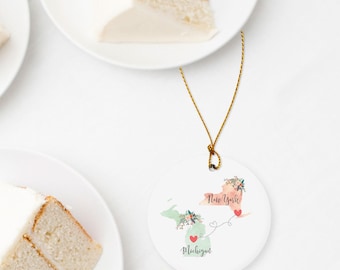 May include: A round white ceramic ornament featuring a watercolor map of New York and Michigan connected by a thin black line with red hearts. The states are decorated with floral accents and the text New York and Michigan in black script.
