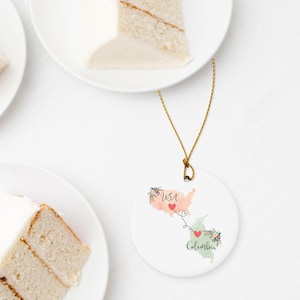 May include: A round white ceramic ornament featuring a watercolor map of the USA in peach and Colombia in light green, connected by a dashed line with two red hearts. The text USA and Colombia is printed in black script with floral accents.
