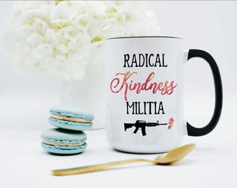 May include: A white ceramic mug with a black handle and rim. The front features the black text RADICAL and MILITIA, with the word Kindness in a pink and orange watercolor script. Below the text is a black rifle graphic firing a small pink flower.