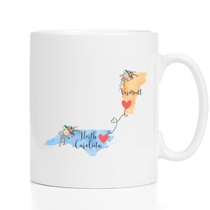 May include: White ceramic coffee mug with a design of the states of Vermont and North Carolina connected by a dotted line with a heart in the middle. The states are outlined in watercolor and have floral accents. The text "Vermont" and "North Carolina" are written on the states.