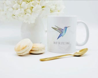 May include: A white ceramic mug featuring a watercolor illustration of a blue and purple hummingbird in flight, with the text "ENJOY THE LITTLE THINGS." printed in black below the bird. The mug has a standard handle and a smooth, glossy finish.