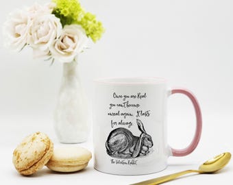 May include: A white ceramic mug with a pink handle and rim, featuring a black ink illustration of a rabbit and the text: Once you are Real you can't become unreal again. It lasts for always. The Velveteen Rabbit is printed below the rabbit.