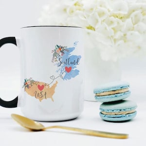 Puede incluir: Una taza de cerámica blanca con asa negra que muestra un mapa de EE. UU. y Escocia conectados por una línea punteada con corazones. Cada mapa está decorado con detalles florales y el texto USA y Scotland en una fuente cursiva.