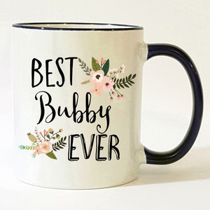 May include: White ceramic mug with a navy blue rim and handle. The mug features a floral design and the text "BEST Bubby EVER" in black.