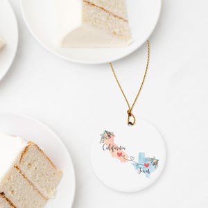 May include: A round white ceramic ornament featuring a peach-colored map of California and a light blue map of Texas. The states are connected by a thin black line with a heart at each end. Floral accents decorate the maps with the text California and Texas.