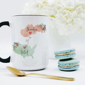 May include: A white ceramic mug with a black handle featuring a watercolor map of Slovakia in peach and the USA in light green. The maps are connected by a dashed line with two red hearts and floral accents. The text reads Slovakia and USA.
