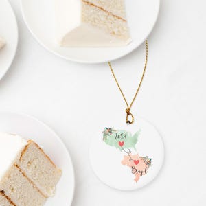 May include: A round white ceramic ornament featuring a watercolor map of the USA in light green and Brazil in light peach. The two countries are connected by a thin black line with two small red hearts. Floral accents decorate the maps.