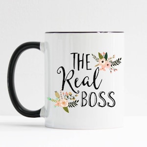 May include: A white ceramic mug with a black handle and rim. The front features the black text THE Real BOSS in a mix of script and block fonts, decorated with small illustrations of pink flowers, green leaves, and brown buds on the sides.