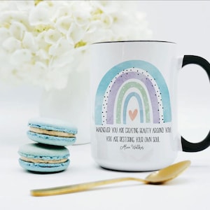 Puede incluir: Una taza de cerámica blanca con asa negra que presenta un gráfico de arcoíris en acuarela en tonos azul, morado y verde con un pequeño corazón color melocotón. El texto dice: "WHENEVER YOU ARE CREATING BEAUTY AROUND YOU, YOU ARE RESTORING YOUR OWN SOUL. Alice Walker."