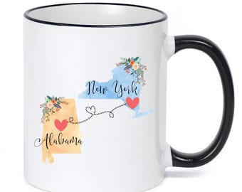 Puede incluir: Una taza de cerámica blanca con borde y asa negros. El diseño presenta un mapa en acuarela de Alabama en color canela y Nueva York en azul, conectados por una línea punteada con corazones. Acentos florales adornan cada estado con el texto.