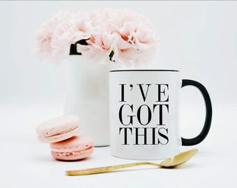 May include: A white ceramic mug with a black handle and rim, featuring the black text I'VE GOT THIS printed on the front. The mug is accompanied by two light pink macarons, a gold-toned spoon, and a white vase filled with light pink flowers.