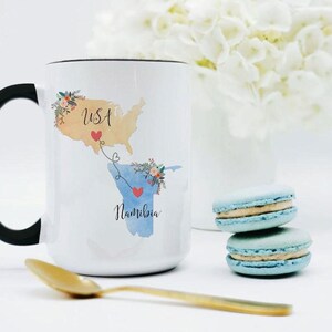 May include: A white ceramic mug with a black handle featuring a watercolor map of the USA in tan and Namibia in blue. The maps are connected by a dotted line with two red hearts and floral accents. The text USA and Namibia is printed on the mug.
