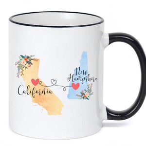 May include: A white ceramic mug with a black rim and handle, featuring a watercolor map of California in yellow and New Hampshire in blue. Two red hearts are connected by a dotted line, with floral accents and the text California and New Hampshire.