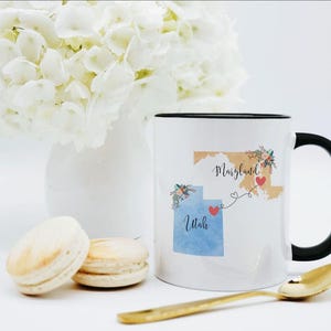May include: A white ceramic mug with a black handle and rim featuring a graphic of the states of Maryland and Utah connected by a dashed line with red hearts. The Maryland shape is tan, and the Utah shape is blue, both decorated with floral accents.
