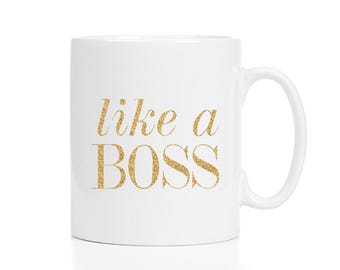 May include: A white ceramic mug featuring the text like a BOSS printed in a gold glitter finish. The word like a is in a cursive script font, while the word BOSS is in a large, bold, capitalized serif font centered on the front of the mug.