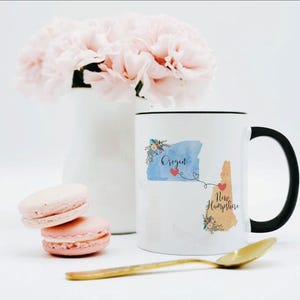 May include: A white ceramic mug with a black handle and rim, featuring watercolor-style maps of Oregon and New Hampshire connected by a dotted line with two red hearts. The text Oregon and New Hampshire is printed in a black script font.