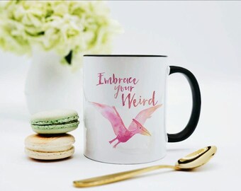 May include: A white ceramic mug with a black handle and rim, featuring a pink watercolor pterodactyl illustration and the text "Embrace your Weird" in a pink script font. The mug is positioned next to two stacked macarons and a gold spoon.
