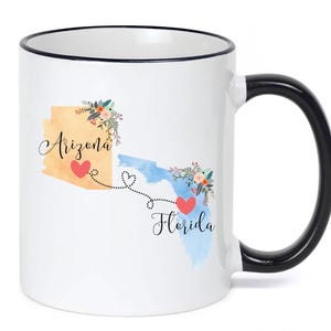 Puede incluir: Una taza de cerámica blanca con borde y asa negros. El diseño presenta un mapa color canela de Arizona y un mapa azul de Florida, conectados por una línea punteada con un corazón. Cada estado tiene un detalle floral y el texto Arizona y Florida.
