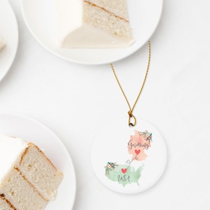 May include: A round white ceramic ornament hanging from a gold string. The ornament features a watercolor map of Germany in peach and the USA in light green, connected by a dashed line with red hearts. Floral accents decorate the maps with text.