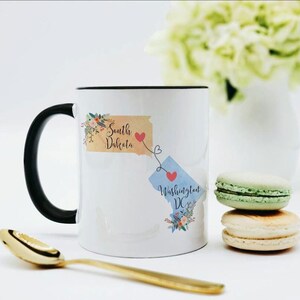Puede incluir: Una taza de cerámica blanca con asa negra que presenta un gráfico de Dakota del Sur en color tostado y Washington DC en azul claro, conectados por una línea punteada con corazones rojos y detalles florales. El texto está impreso en la taza.