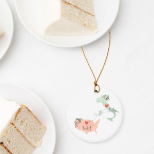 May include: A round white ceramic ornament featuring a map of the USA in peach and a map of Italy in light green. Both maps are decorated with floral accents and red hearts connected by a thin line. The text USA and Italy is printed below.