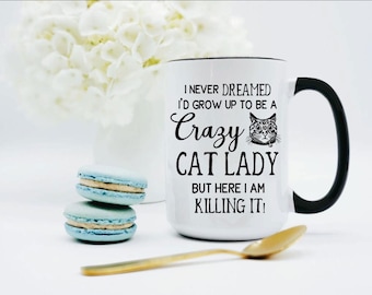 May include: A white ceramic mug with a black handle and interior, featuring the black text I NEVER DREAMED I'D GROW UP TO BE A Crazy CAT LADY BUT HERE I AM KILLING IT! along with a small black and white illustration of a cat face.