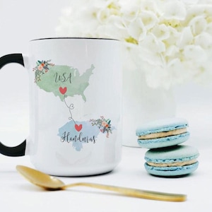 May include: A white ceramic mug with a black handle featuring a watercolor map of the USA and Honduras connected by a dotted line with red hearts. The text USA and Honduras is printed on the maps, which are decorated with small floral accents.