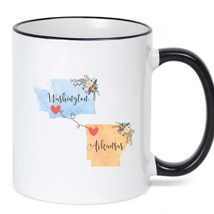 May include: White ceramic mug with a black rim and handle. The mug features a watercolor design of the states of Washington and Arkansas with hearts and floral accents. The text "Washington" and "Arkansas" is printed on the mug.
