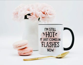 May include: A white ceramic mug with a black handle and rim, featuring the black text I'M STILL HOT IT JUST COMES IN FLASHES NOW printed over a soft pink watercolor design. The mug is positioned next to two pink macarons and a gold spoon.