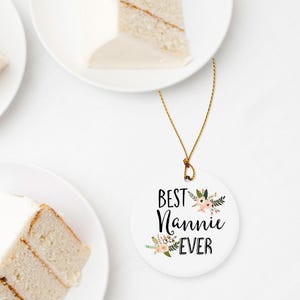 Puede incluir: Un adorno redondo de cerámica blanca que cuelga de un cordón dorado. El adorno presenta el texto negro "BEST Nannie EVER" en una mezcla de fuentes cursivas y de bloque, decorado con pequeñas ilustraciones de flores rosas y hojas verdes.