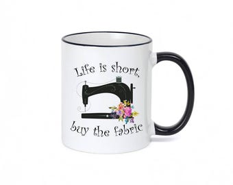 May include: A white ceramic mug with a black rim and handle. The front features a black illustration of a sewing machine adorned with a colorful floral arrangement. The text reads, Life is short, buy the fabric, in a black cursive script font.