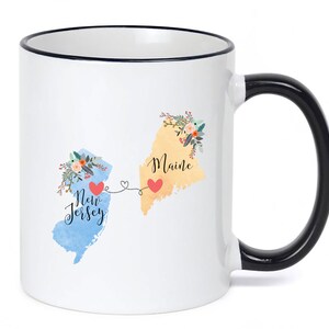 May include: White ceramic coffee mug with a black rim and handle. The mug features a design of the states of Maine and New Jersey with floral accents and the words "Maine" and "New Jersey".