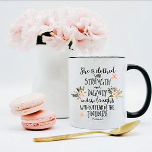 May include: A white ceramic mug with a black handle and rim, featuring the black printed text 'She is clothed with STRENGTH and DIGNITY and she laughs WITHOUT FEAR OF THE FUTURE Proverbs 31:25' accented with small pink and green floral illustrations.