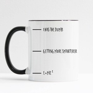 May include: White ceramic mug with a black handle and black rim. The mug has three lines of text: "I has the dumb", "Getting more smarterer", and "E=MC^2".