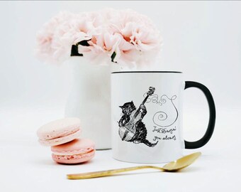 May include: A white ceramic mug with a black handle and rim, featuring a black ink illustration of a cat playing a cello. The text "Just stringin' you along" is printed in a cursive script next to the cat. The mug has a smooth, glossy finish.