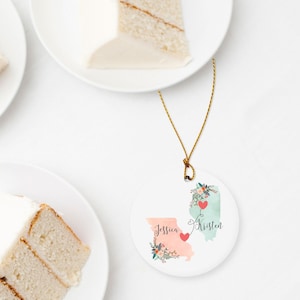 May include: A round white ceramic ornament hanging from a gold string. The ornament features a peach-colored map of Missouri and a light green map of Illinois, each decorated with floral accents, red hearts, and the names Jessica and Kristen.