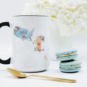May include: A white ceramic mug with a black handle featuring a blue map of the USA and a tan map of Bahrain connected by a dashed line with red hearts. Floral accents decorate the top of each map, and the text USA and Bahrain is printed below.