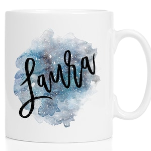 Puede incluir: Una taza de cerámica blanca con un diseño de acuarela en tonos azules y grises con motas blancas que recuerdan a un cielo estrellado. El nombre Laura está impreso en el centro del gráfico de acuarela en una letra cursiva negra y negrita.