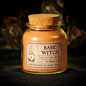 May include: A glass jar candle with a cork lid. The candle is filled with orange wax and has a label that reads "BASIC WITCH". The label also lists the scent as "Pumpkin Spice Latte, Milk Foam" and "Coco-Beeswax Candle".