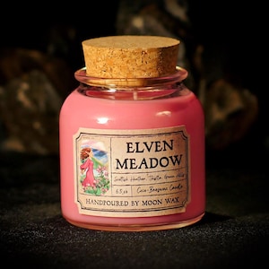 May include: A pink candle in a glass jar with a cork lid. The label reads "Elven Meadow" and includes an illustration of a woman in a pink dress. The candle is hand-poured and contains 6.5 oz of wax.
