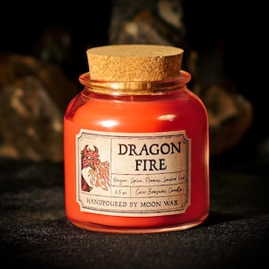 May include: A small, round glass jar candle with a cork stopper. The candle wax is a vibrant orange-red color. A label on the front reads "DRAGON FIRE" with a dragon illustration. The label also lists ingredients and the text "HANDPOURED BY MOON WAX".